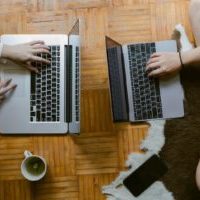 From above of crop unrecognisable female friends typing while studying on laptops remotely and enjoying tea on floor in cozy living room